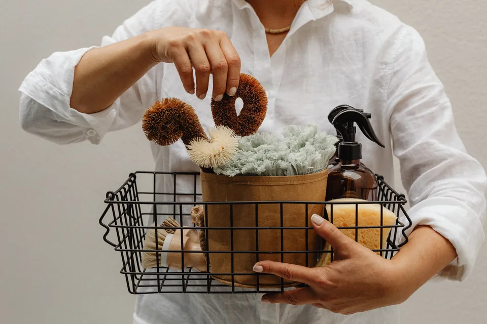 Cropped hands choosing a scrub brush from a black wire basket of cloths, sponges and an amber spray bottle.